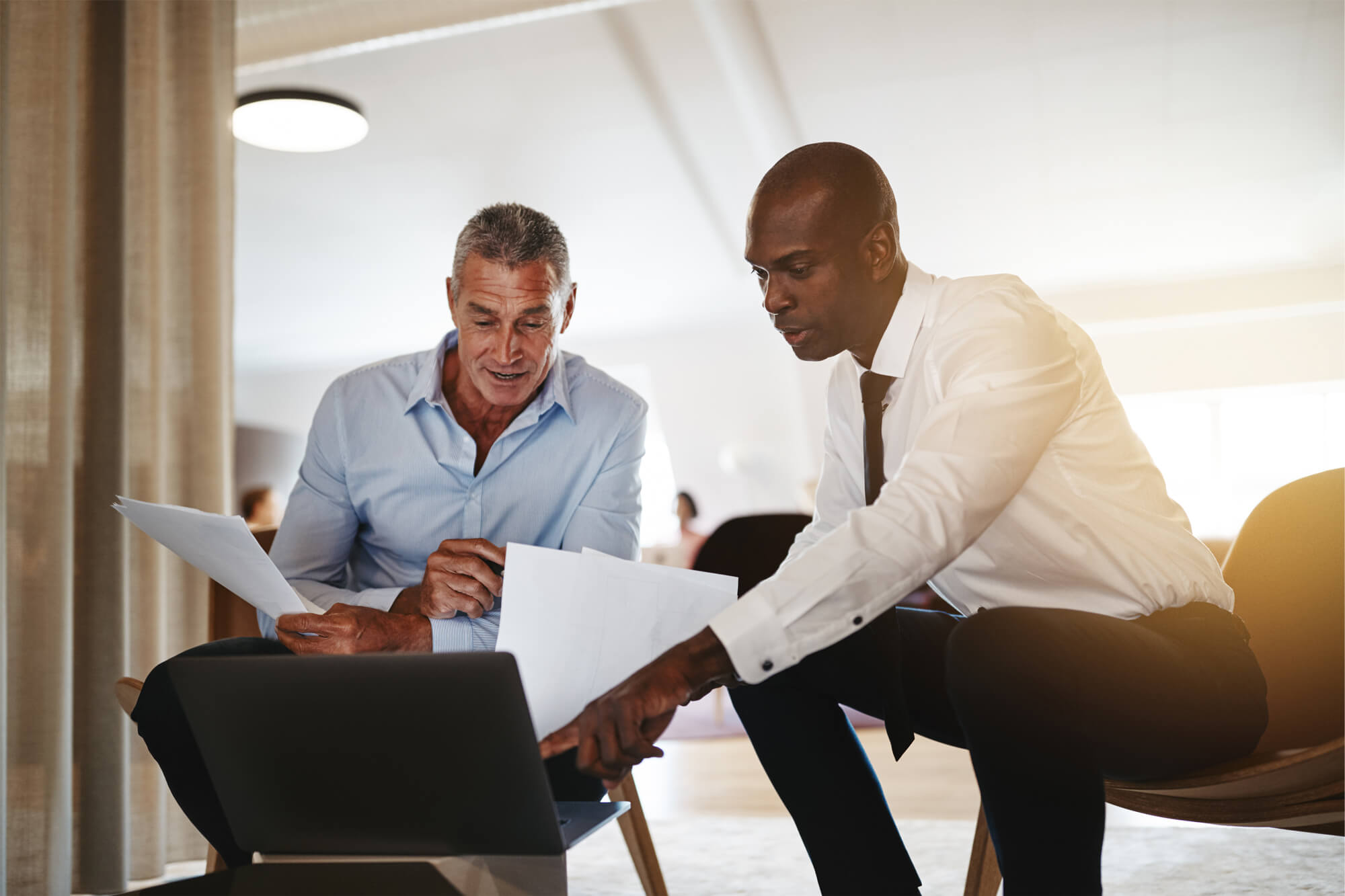 Two diverse businessmen sitting together in a modern office discussing paperwork and working on a laptop